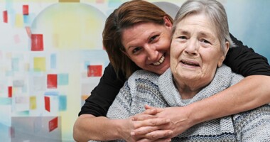 Eine junge Frau umarmt eine alte Frau, beide lächeln in die Kamera.